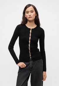 Črna rebrasta majica z dolgimi rokavi, ki ima okroglo izrez, z zaklepom na ključavnico in bisernimi detajli ter obliko, ki se prilega telesu.