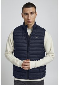 Navyblaue Steppweste mit hohem Kragen, Frontreißverschluss und horizontalem Steppmuster. Getragen über einem cremefarbenen Langarmshirt.