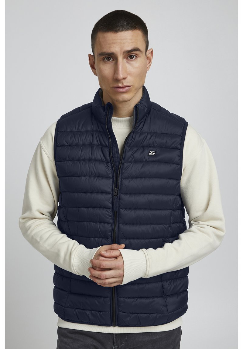 Navyblaue Steppweste mit hohem Kragen, Frontreißverschluss und horizontalem Steppmuster. Getragen über einem cremefarbenen Langarmshirt.