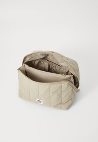 Beige quiltet veske med glidelåsåpning, som har et romslig innvendig rom og en ekstra glidelåslomme. Mykt, teksturert stoff.