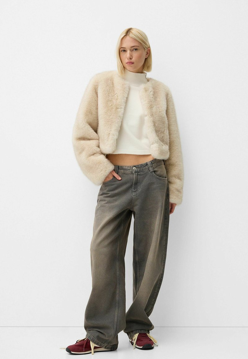 Giovane donna che indossa una giacca in pelliccia sintetica beige, un maglione bianco corto, jeans grigi sbiaditi larghi e sneakers bordeaux, in piedi su uno sfondo bianco.