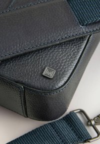 Prostokątna torebka ze skóry w kolorze granatowym z teksturowanym wykończeniem, niebieskim przeszyciem i metalowym akcentem z logo. Zawiera odpinany pleciony pasek.