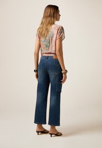 Pantaloni cargo in denim con gamba larga, lavaggio scuro e molte tasche, abbinati a una maglietta a maniche corte a fantasia nei colori pastello.