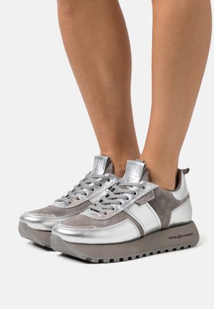 Paire de jambes portant des baskets à lacets gris et argent avec des semelles épaisses à crampons sur un fond clair uni.