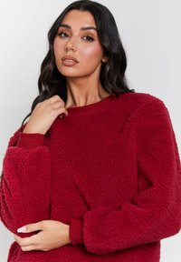 Röd fleece-tröja med en fluffig textur, rund halsringning och ribbade ärmar. Materialet är mjukt och har en enfärgad yta utan mönster.