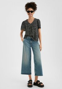 Schwarze Bluse mit Polka-Dots und kurzen Ärmeln, kombiniert mit weit geschnittenen, hellblauen Jeans und schwarzen Plateausandalen. Glatte Texturen, lässige Passform.
