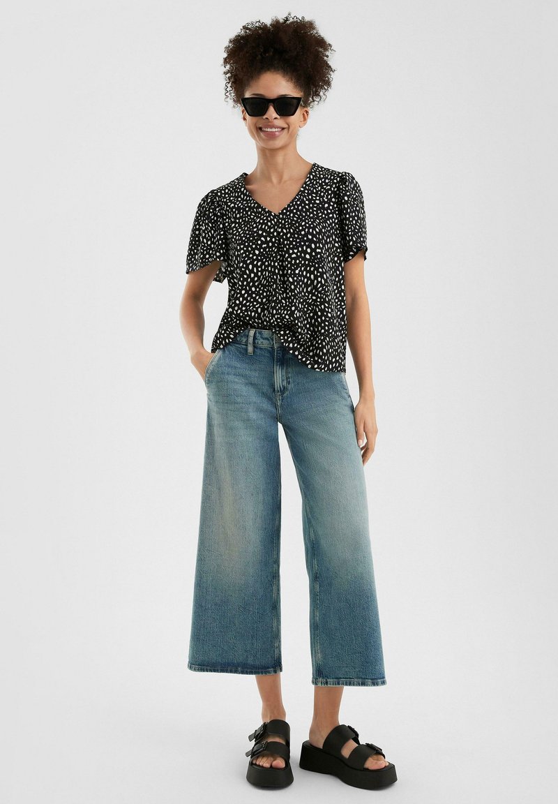 Schwarze Bluse mit Polka-Dots und kurzen Ärmeln, kombiniert mit weit geschnittenen, hellblauen Jeans und schwarzen Plateausandalen. Glatte Texturen, lässige Passform.
