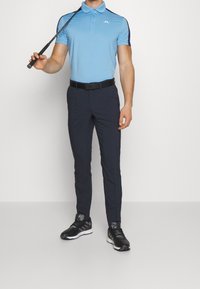 Ljusblå pikétröja med marinblå detaljer, kombinerad med mörkblå byxor och svarta sportskor. Tröjan har en logga på bröstet.