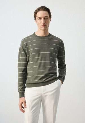 Hombre con suéter de rayas finas color verde oliva y pantalones blancos, de pie con una mano en el bolsillo, fondo neutral.