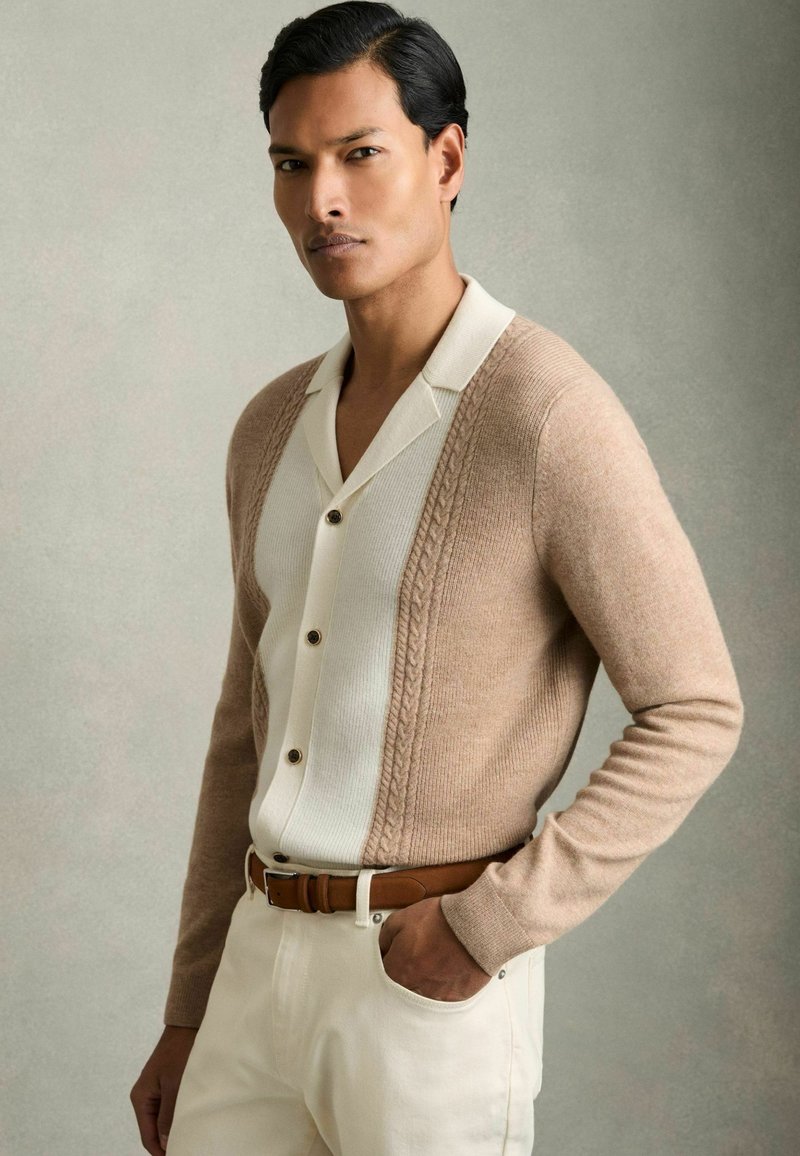 Gilet en tricot beige avec un motif en cable, associé à une chemise blanche à boutons avec un col doux, une ceinture marron et un pantalon de couleur claire.