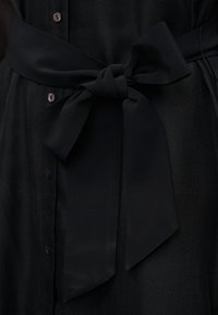 Μαύρη υφασμάτινη ζώνη δεμένη σε φιόγκο γύρω από ένα υφασμάτινο μαύρο ένδυμα με υφή και κουμπιά.