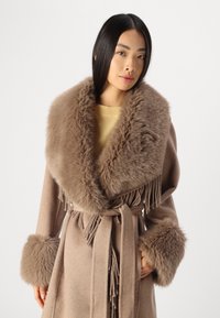 Μπεζ μάλλινο παλτό με μεγάλο κολάρο και μανσέτες από ψεύτικη γούνα. Διαθέτει ζώνη στη μέση και κρόσσια στο μπροστινό μέρος. Υφή μαλακή και με υφή.