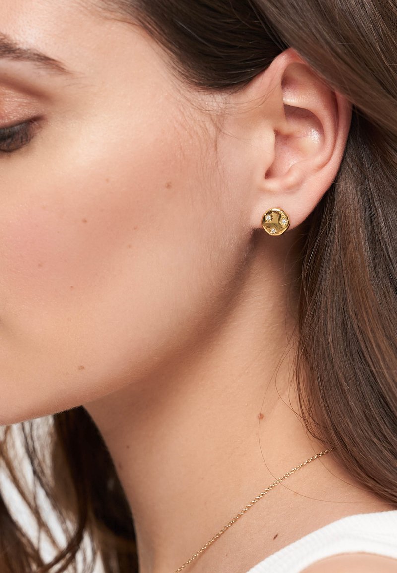 Orecchini a bottone in oro con un design rotondo e testurizzato, caratterizzati da tre piccoli diamanti incastonati. Indossati all'orecchio di un modello.