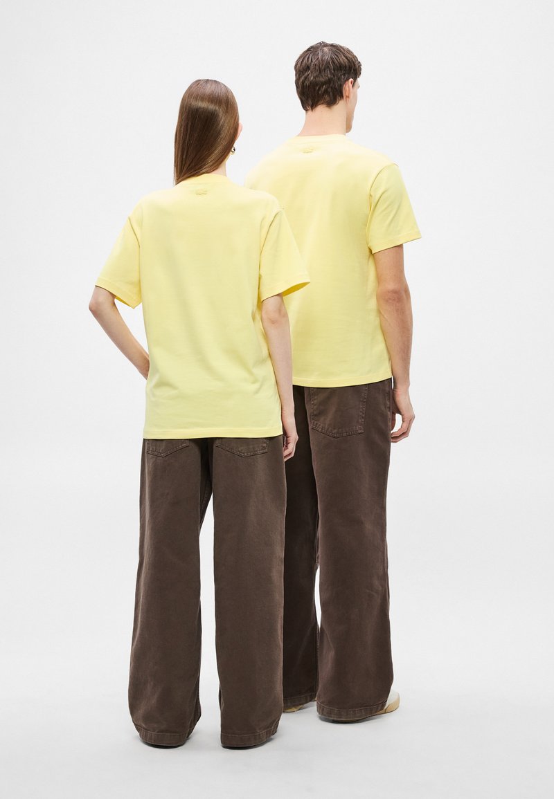 T-shirt gialle in cotone con maniche corte, abbinate a pantaloni marroni ampi. Design semplice senza grafiche o loghi visibili.