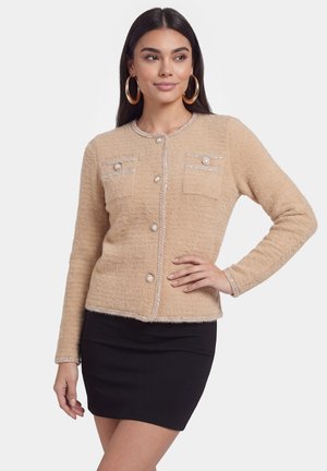 Donna che indossa un cardigan beige effetto texture con bottoni e tasche decorative e una gonna nera aderente, in posa con una mano sui fianchi.