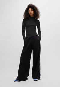 Zwarte geribde turtleneck trui gecombineerd met wijde zwarte broeken met witte zijstreep, gedragen met blauw-witte sneakers.