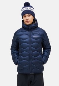 Navy blue puffer jacket with quilted wave pattern, high collar, front zipper, and adjustable hood. Worn with a matching navy beanie.