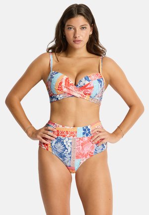 Femme portant un bikini taille haute coloré avec des motifs floraux et géométriques, debout, les mains sur les hanches, sur un fond uni.