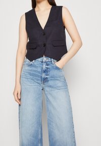 Marineblaue ärmellose Weste mit V-Ausschnitt, zwei vorderen Taschen und schwarzen Knöpfen, kombiniert mit hellblauen weiten Jeans. Glatte Stoffstruktur.