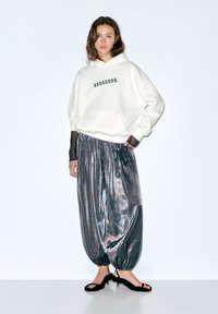 Felpa bianca con motivo a stelle nere, abbinata a pantaloni larghi grigio lucido. Modello indossa tacchi neri con cinturini. Sfondo semplice.