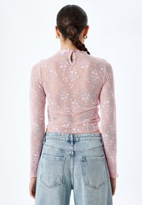 Rosa Spitzenoberteil mit langen Ärmeln, Blumenmuster, Rüschenausschnitt und einem kleinen Schlüssellochdetail auf der Rückseite, kombiniert mit hellblauen Denim-Jeans.
