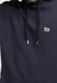 Mörkblå hoodie i bomullsblandning, med en liten vit "Lee"-logotyp, snörning med metallspetsar och en kenguruficka.