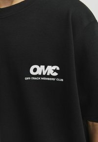 Juodas medvilnės marškinėliai su apvaliu kaklu ir trumpomis rankovėmis. Ant jų pavaizduotas baltas spausdintas logotipas ir tekstas "OMC OFF-TRACK MEMBERS' CLUB."