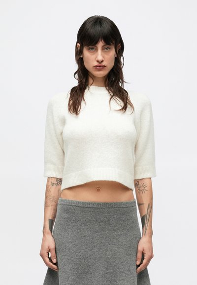 Pull court en maille crème coupé, avec des manches courtes, un col côtelé et une texture douce. Associé à une jupe grise, laissant entrevoir la peau au niveau de la taille.