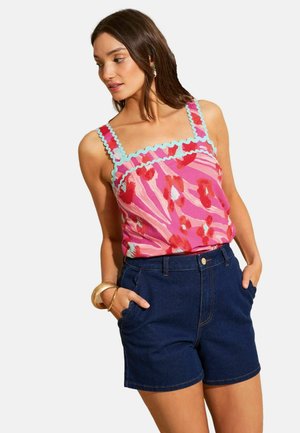 Kvinde iført pink blomsterprintet ærmeløs top med blå kant og mørkeblå denimshorts, hænderne i lommerne, kigger mod venstre.