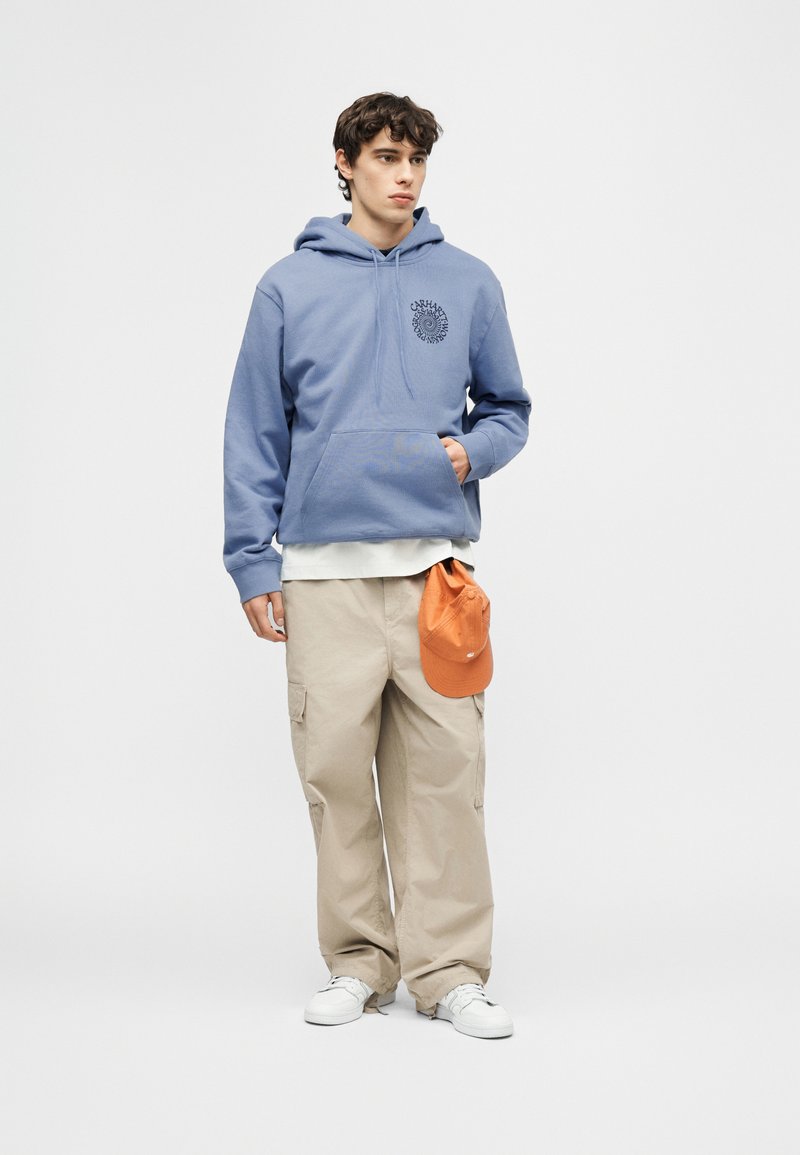Joven con sudadera azul con logo, pantalones cargo beige sosteniendo una gorra naranja y zapatillas blancas, de pie frente a un fondo liso.