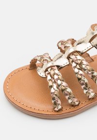 Sandale à bride tressée en or avec des accents métalliques, une semelle en cuir beige et une texture lisse. Le design comprend trois sections tressées pour un bon maintien.