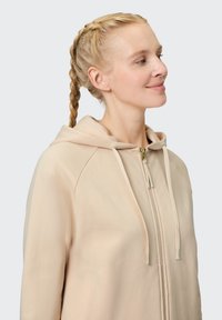 Peach-colored zip-up hoodie with a front pocket, drawn hoodie strings, and smooth fabric texture, worn by a person with a side profile.