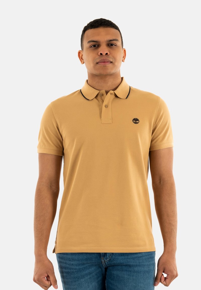 Chemise polo beige avec un col et des manches courtes, présentant un col noir bordé et un petit logo sur la poitrine. Portée avec un jean bleu.
