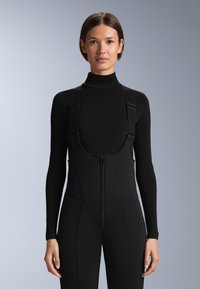 Žena oblečená v černém topu s vysokým límcem a dlouhými rukávy a černých laclích na zip s nastavitelnými ramenními popruhy, stojící na šedém pozadí.
