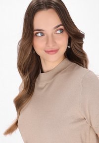Lys beige sweater med høj hals og korte ærmer, der har en blød tekstur og en subtil glans. Modellen har langt, bølget hår.