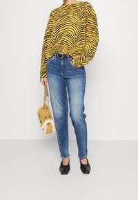 Persona vistiendo un suéter a rayas de tigre amarillo y negro, jeans azules con cinturón negro, zapatos negros, llevando un bolso acolchado amarillo con ribete de piel.