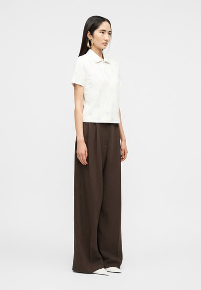 Femme aux longs cheveux foncés porte une chemise blanche à manches courtes et col, de grandes boucles d'oreilles dorées, un pantalon large marron et des chaussures blanches pointues, debout de profil.