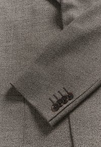 Gros plan sur une manche de veste en tweed gris avec quatre boutons foncés cousus près du poignet.