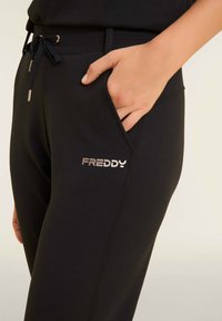 Pantalons de survêtement noirs avec poche latérale, taille élastique, cordon de serrage réglable et détail de logo métallique indiquant "FREDDY." Texture lisse.