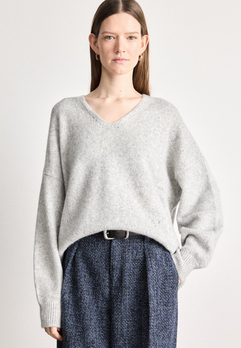 Sweater solto de cor cinza claro com decote em V e punhos canelados, combinado com uma saia xadrez texturizada azul escura e um cinto preto.