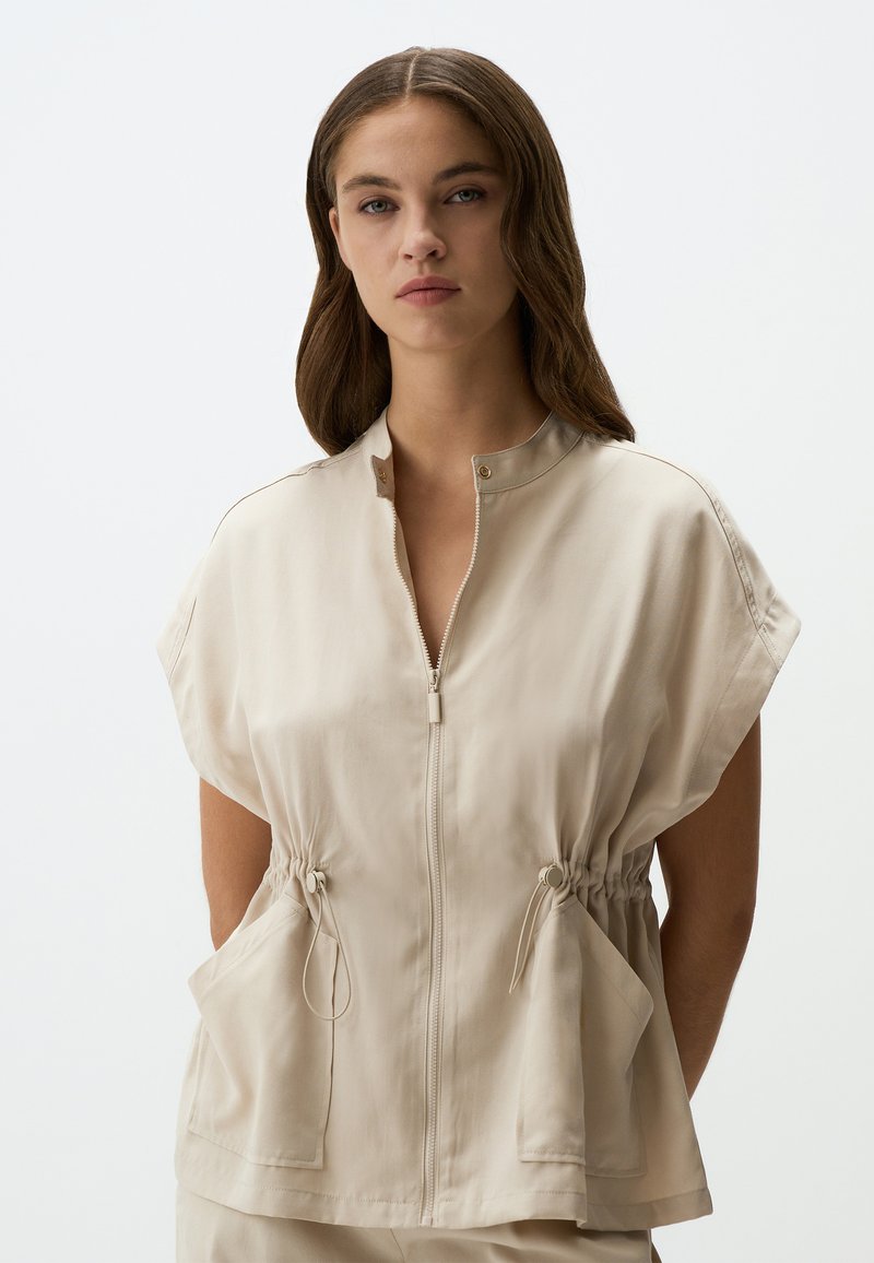 Beige, kurzärmliges Oberteil mit Reißverschluss vorne, Stehkragen und elastischem Zugband in der Taille. Besitzt zwei Vordertaschen. Glatte Textur.