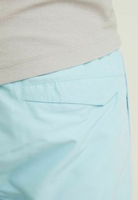 Pantalon bleu clair avec une poche arrière et un tissu beige texturé légèrement rentré au-dessus de la taille, sur un fond clair uni.