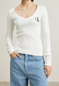 Vit långärmad ribbad topp med djup V-ringning, med en liten mörk logotyp på bröstet, ihop med ljusa denimjeans.