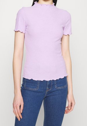 Femme portant un haut moulant côtelé lavande à manches courtes avec des bords en forme de laitue, associé à un jean bleu taille haute avec des poches avant.
