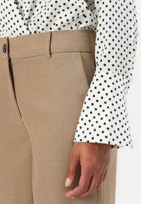 Lys brune tailormakkede bukser med flat foran, parret med en hvid bluse med sorte polka prikker. Glat stof og raffineret snit.