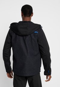 Chaqueta con capucha en negro y antracita, que cuenta con una textura suave, puños elásticos y un detalle del logo azul en el hombro izquierdo de la espalda.