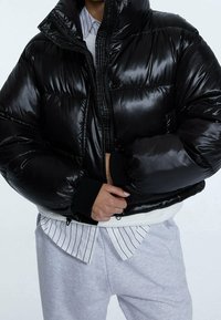 Giacca a vento nera con finitura lucida, colletto alto e polsini elasticizzati. Presenta una zip frontale e orlo a coste, indossata sopra una camicia a righe.
