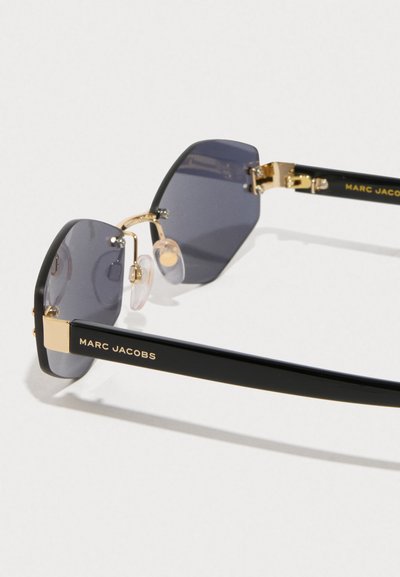 Μαύρα γυαλιά ηλίου με εξαγωνικούς, σκούρους γκρίζους φακούς. Χρυσές λεπτομέρειες σε μεταλλικό σκελετό και διάφανα στηρίγματα μύτης. Λογότυπο Marc Jacobs στους βραχίονες.