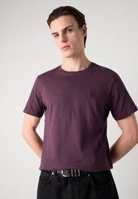 Tricou cu mânecă scurtă, decupaj rotund, din bumbac de culoare mov închis, cu o mică etichetă texturată pe piept. Asortat cu blugi negri și curea.