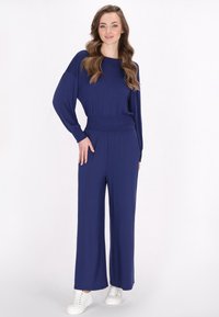 Marineblauw jumpsuit met lange mouwen, smocktail, wijd uitlopende pijpen en een soepele, rekbare stof. Gecombineerd met witte sneakers.